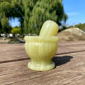 May include: A light green stone mortar and pestle set featuring a smooth, polished finish with subtle, vertical white banding throughout the cylindrical body and the rounded, tapered pestle handle.