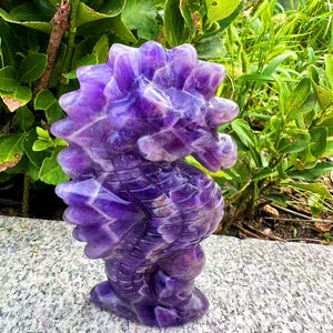 May include: A carved amethyst seahorse sculpture. The seahorse is a deep purple color with lighter purple and white veining throughout. The sculpture is standing on a gray stone surface, with green foliage in the background.