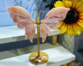 May include: A pink and purple butterfly sculpture made of stone, mounted on a gold-colored metal stand. The butterfly has detailed wing patterns and is displayed on a white shelf. The background includes sunflowers and greenery.