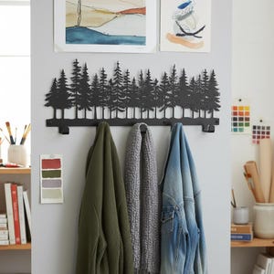 May include: A black metal wall-mounted coat rack featuring a forest silhouette design. The rack has multiple hooks and is holding a green sweater, a gray scarf, and a blue denim jacket. Two framed paintings hang above the rack.