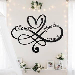 May include: Black metal wall art featuring the names "Clint" and "Claudia" intertwined with an infinity symbol and a heart. The date "8.11.2025" is also included. The artwork is displayed on a white wall, with floral arrangements and framed photos below.