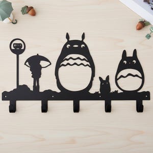 Może przedstawiać: Czarny, metalowy wieszak ścienny z sylwetkami postaci z filmu animowanego "M&oacute;j sąsiad Totoro". Wz&oacute;r obejmuje znak przystanku autobusowego, osobę z parasolem i trzy postacie Totoro, z czterema haczykami do wieszania przedmiot&oacute;w.