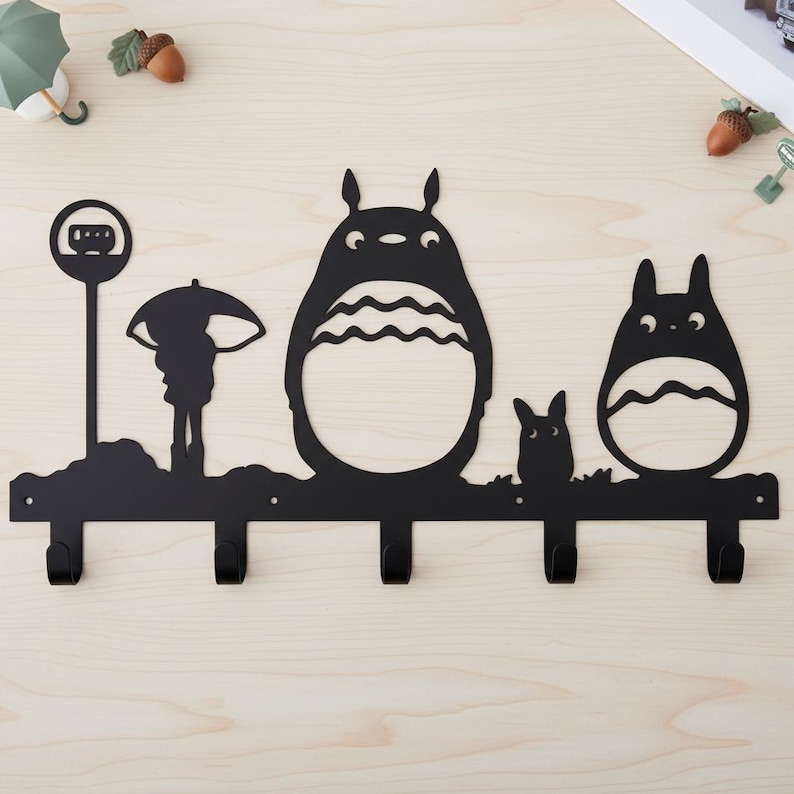 Może przedstawiać: Czarny, metalowy wieszak ścienny z sylwetkami postaci z filmu animowanego "M&oacute;j sąsiad Totoro". Wz&oacute;r obejmuje znak przystanku autobusowego, osobę z parasolem i trzy postacie Totoro, z czterema haczykami do wieszania przedmiot&oacute;w.