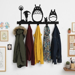 May include: Black metal wall-mounted coat rack featuring a silhouette of characters from a popular animated film. The rack has five hooks and is holding a variety of coats and scarves. The design includes a bus stop, an umbrella, and three cartoon figures.