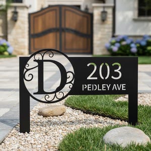 Könnte beinhalten: Schwarzes Metall-Adressschild mit der Nummer 203 und dem Straßennamen "PEDLEY AVE" in Weiß. Das Schild hat ein dekoratives kreisförmiges Design mit dem Buchstaben "D". Das Schild ist auf zwei Pfosten montiert und in einem Beet aus kleinen Steinen und Gras platziert.