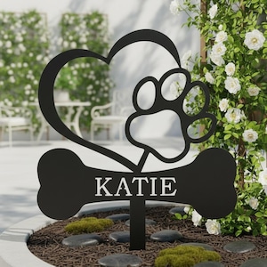 May include: Black metal garden stake featuring a heart, paw print, and bone design. The bone displays the name "KATIE" in white lettering. Ideal for pet owners, this decorative stake adds a personalized touch to any garden or outdoor space.