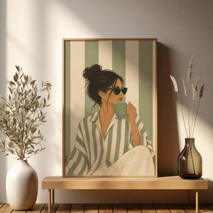Könnte beinhalten: Gerahmter Kunstdruck mit einer Frau mit Sonnenbrille und einer Tasse. Sie trägt ein gestreiftes Hemd und eine weiße Hose vor einem grün-weiß gestreiften Hintergrund. Das Kunstwerk steht auf einer Holzbank mit dekorativen Vasen.