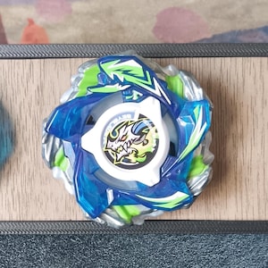 Puede incluir: Tres coloridas peonzas exhibidas sobre una superficie de madera dentro de un marco negro. Las peonzas presentan detalles en azul, verde y blanco. Las peonzas están diseñadas con detalles y patrones intrincados, mostrando una variedad de formas y texturas.