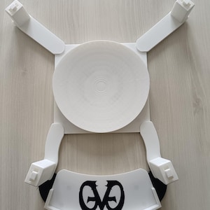 Puede incluir: Un dispositivo blanco impreso en 3D con un centro circular y cuatro brazos que se extienden, cada uno terminando en un bloque rectangular. Una pieza blanca curva con detalles en negro y las letras "EVO" en la parte inferior. El dispositivo está sobre una superficie clara de vetas de madera.