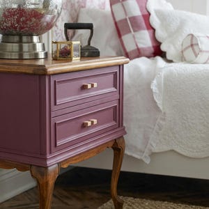 Puede incluir: Una mesita de noche color ciruela con dos cajones y tiradores de madera. La mesita tiene una tapa de madera y patas curvas. Una lámpara decorativa de cristal y una pequeña caja dorada están encima. Un cojín a cuadros rojos y blancos está en el fondo.