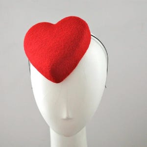 May include: A bright red heart-shaped headpiece. The heart is made of a textured material and is attached to a thin black headband. The headpiece is displayed on a white mannequin head against a plain background. This accessory is suitable for special occasions.