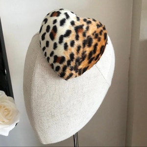 May include: A heart-shaped fascinator with a leopard print pattern in shades of brown, black, and cream. The hat is mounted on a neutral-colored head form, showcasing the design's texture and shape. A unique accessory.