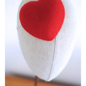 May include: A red heart-shaped decoration is affixed to a mannequin head. The heart is a vibrant red color and appears to be made of a soft, textured material. The mannequin head is a neutral beige color.