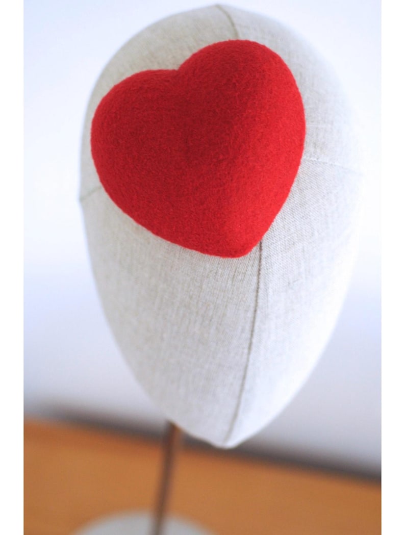 May include: A red heart-shaped decoration is affixed to a mannequin head. The heart is a vibrant red color and appears to be made of a soft, textured material. The mannequin head is a neutral beige color.