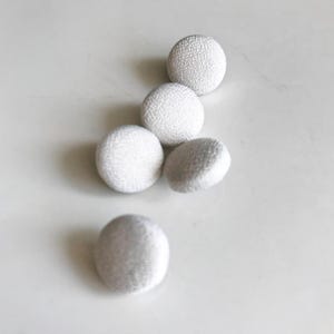 Op de afbeelding: Vijf kleine, ronde, met stof bedekte knopen in verschillende posities. De knopen hebben een lichte, neutrale kleur met een getextureerd oppervlak. De knopen zijn waarschijnlijk voor naai- of knutselprojecten.