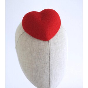 May include: A red heart-shaped headpiece atop a mannequin head. The heart is a vibrant red and appears to be made of a textured material. The mannequin head is a neutral beige color, and the background is plain white.