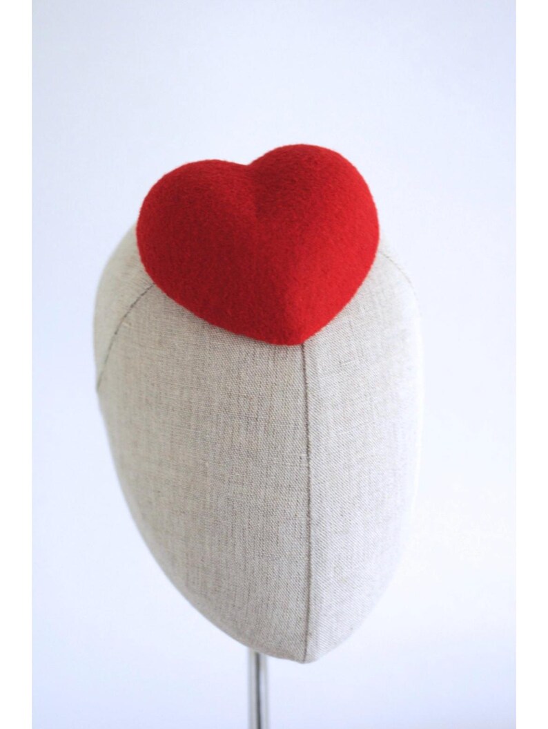 May include: A red heart-shaped headpiece atop a mannequin head. The heart is a vibrant red and appears to be made of a textured material. The mannequin head is a neutral beige color, and the background is plain white.