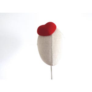 May include: A red heart-shaped hat is displayed on a mannequin head. The hat is made of a felt-like material and sits atop the head form. The mannequin head is covered in a light beige fabric. The background is plain white.
