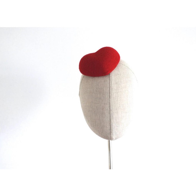 May include: A red heart-shaped hat is displayed on a mannequin head. The hat is made of a felt-like material and sits atop the head form. The mannequin head is covered in a light beige fabric. The background is plain white.