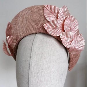 May include: A blush-colored hat with a rounded crown and a decorative arrangement of pink fabric leaves. The hat is made of a woven material, and the leaves have a textured, metallic finish. This headwear is suitable for special occasions.