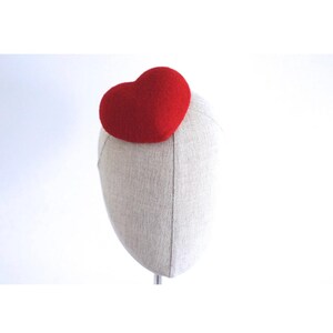 May include: A red heart-shaped headpiece, possibly a fascinator, sits atop a mannequin head. The heart is a vibrant red, and the mannequin head is covered in a light-colored fabric. The background is plain white.