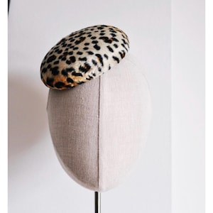 May include: A round hat with a leopard print pattern, featuring black and brown spots on a cream background. The hat is displayed on a mannequin head. The hat's design is a classic, stylish accessory.