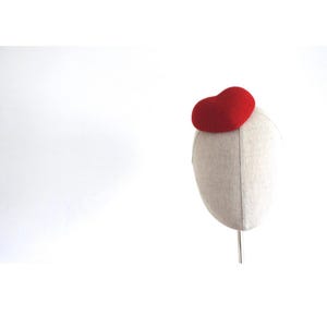 May include: A red felt pillbox hat atop a mannequin head. The hat is a solid, rounded shape. The mannequin head is covered in a light beige fabric. The background is plain white.