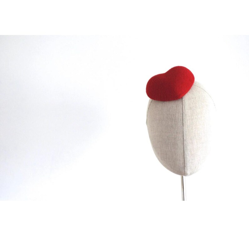 May include: A red felt pillbox hat atop a mannequin head. The hat is a solid, rounded shape. The mannequin head is covered in a light beige fabric. The background is plain white.