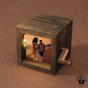 以下が含まれることがあります： ビーチにいるカップルの写真が入った木製のオルゴール。立方体型の箱には「Together wherever」の文字が刻まれています。小さなクランクが側面にあります。箱は小さなライトで照らされています。