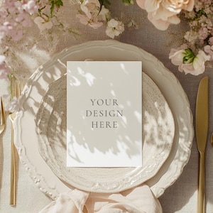 May include: A table setting with a white card that reads "YOUR DESIGN HERE" on a white plate. Gold-coloured fork, knife, and spoon are arranged on the table. The table is decorated with pink flowers and a beige napkin.
