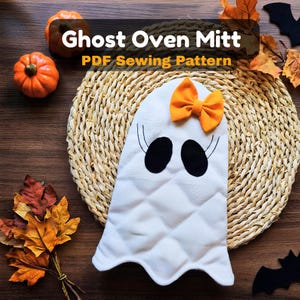 Könnte beinhalten: Ein weißer, gesteppter Ofenhandschuh in Geisterform mit schwarzen ovalen Augen, geschwungenen Wimpern und einer orangefarbenen Stoffschleife. Der Text Ghost Oven Mitt PDF Sewing Pattern ist sichtbar. Der Handschuh liegt auf einer runden Matte.