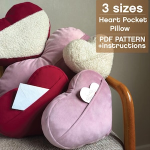 May include: Heart-shaped pillows in shades of red and pink, some with a soft, textured surface. One pillow has a pocket with a heart-shaped card. The image includes text that reads "3 sizes Heart Pocket Pillow PDF PATTERN +instructions."
