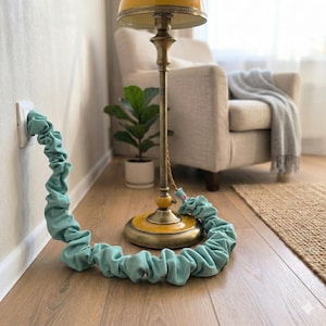 May include: A teal fabric cord cover is connected to a wall outlet and a brass floor lamp with a yellow lampshade. The cord cover is on a wooden floor. A potted plant and a beige armchair are in the background.