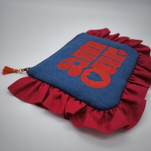 May include: A denim blue clutch with red embroidered text and a red ruffled border. The clutch has a small orange tassel attached to the zipper. The text on the clutch is in a bold, stylized font.