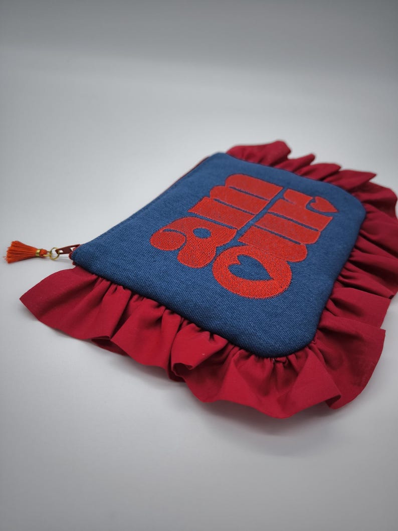 May include: A denim blue clutch with red embroidered text and a red ruffled border. The clutch has a small orange tassel attached to the zipper. The text on the clutch is in a bold, stylized font.