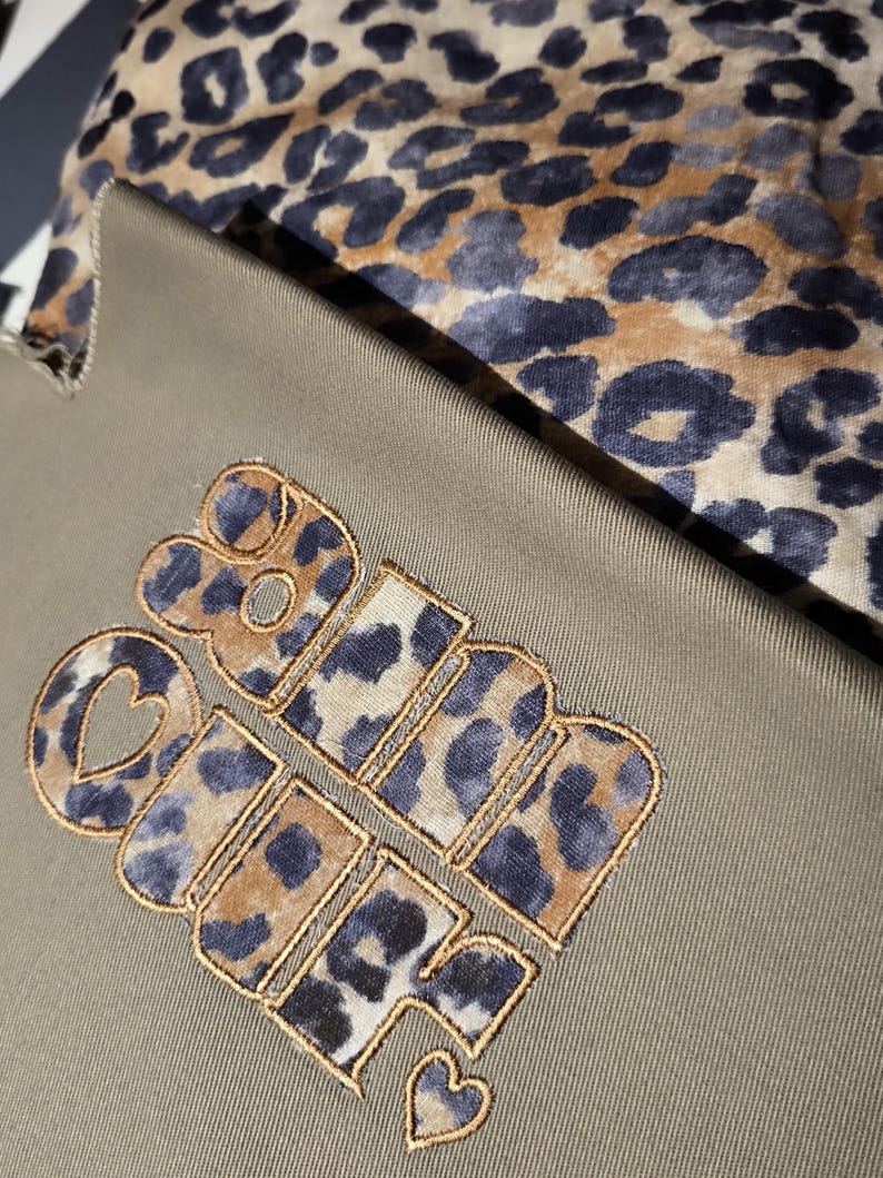 May include: Close-up of a khaki-colored fabric with the embroidered words "Girl Power" in a leopard print design. A leopard print fabric is visible in the background.