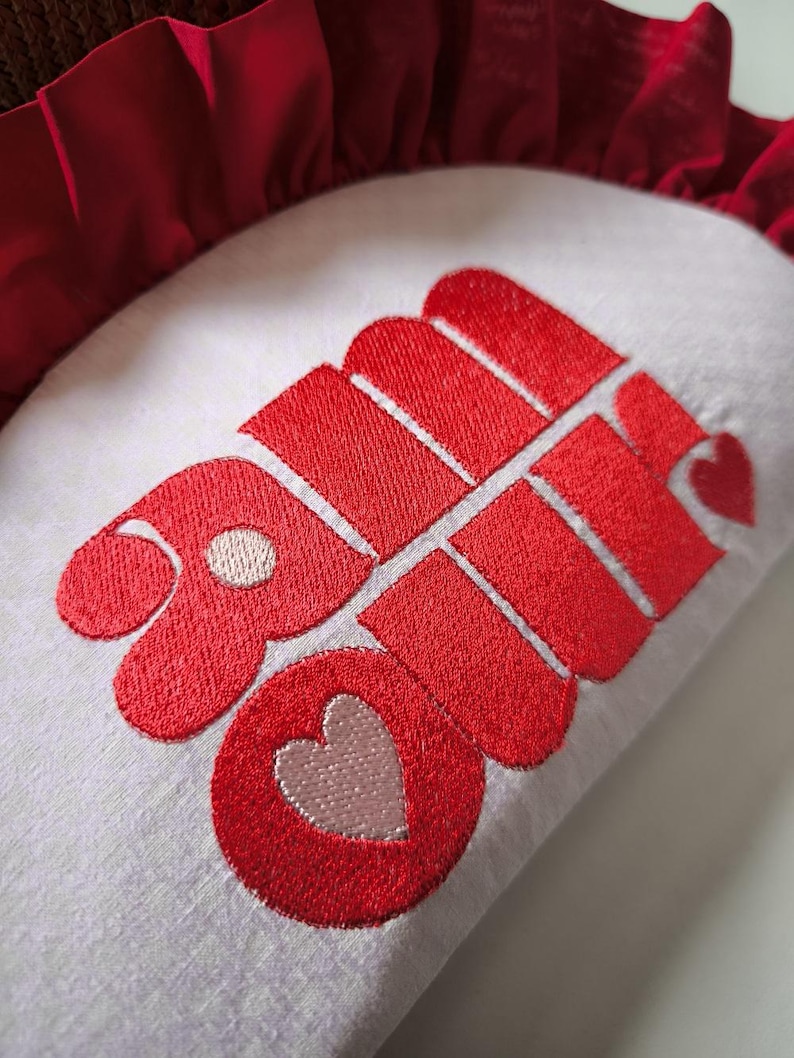 May include: A white pillow with a red embroidered design. The design features the word "Love" in a stylized font, with a heart incorporated into the design. The pillow has a red ruffled border.