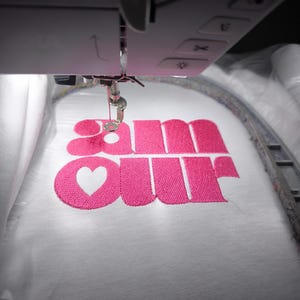 Peut inclure: Gros plan d'une machine à broder brodant le mot "amour" en rose vif sur un tissu blanc. Le "o" est remplacé par un cœur. L'aiguille et le cadre de la machine sont visibles.