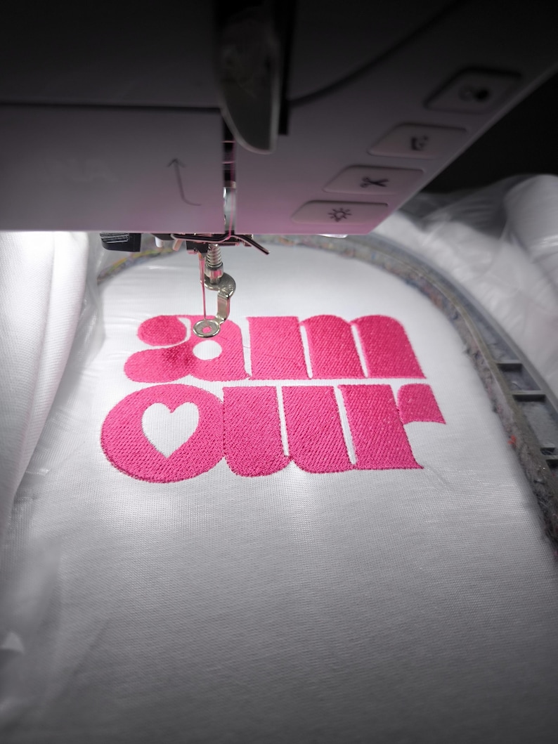 May include: Close-up of an embroidery machine stitching the word "amour" in bright pink thread onto a white fabric. The "o" is replaced with a heart shape. The machine's needle and frame are visible.