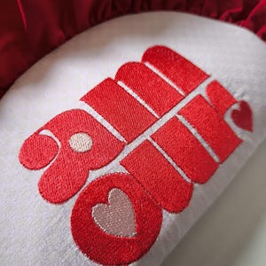 Peut inclure: Un coussin blanc avec un motif brodé rouge. Le motif présente le mot "Love" dans une police stylisée, avec un cœur intégré dans le design. Le coussin a une bordure rouge à volants.