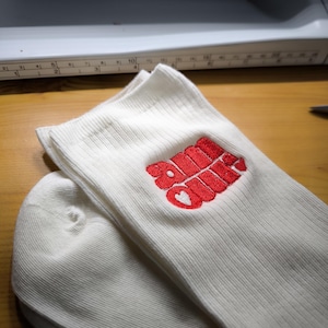 May include: White ribbed socks with a red embroidered design that includes the word "Soul" and a heart. The socks are folded and stacked on a wooden surface, with a sewing machine visible in the background.