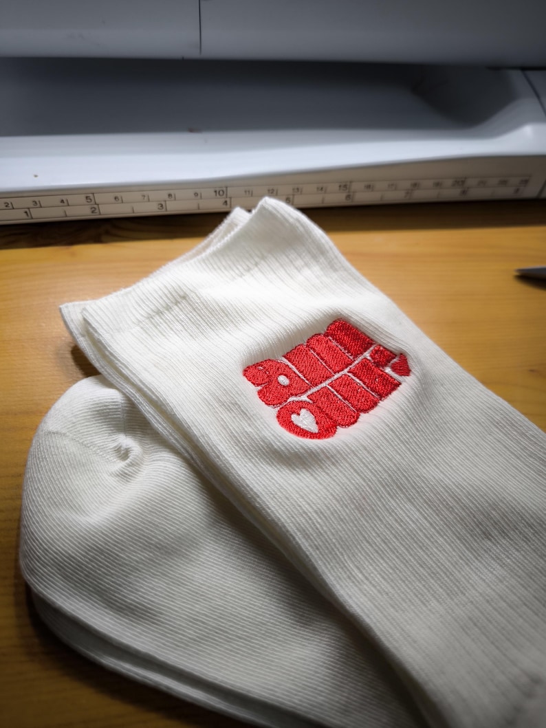 May include: White ribbed socks with a red embroidered design that includes the word "Soul" and a heart. The socks are folded and stacked on a wooden surface, with a sewing machine visible in the background.