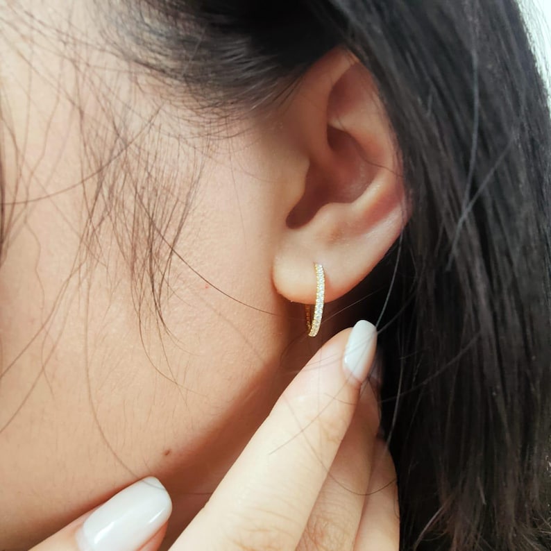May include: A close-up of a gold hoop earring with small, sparkling stones. The earring is worn on the ear. The earring is a simple, elegant design. The background is blurred, focusing attention on the jewelry.