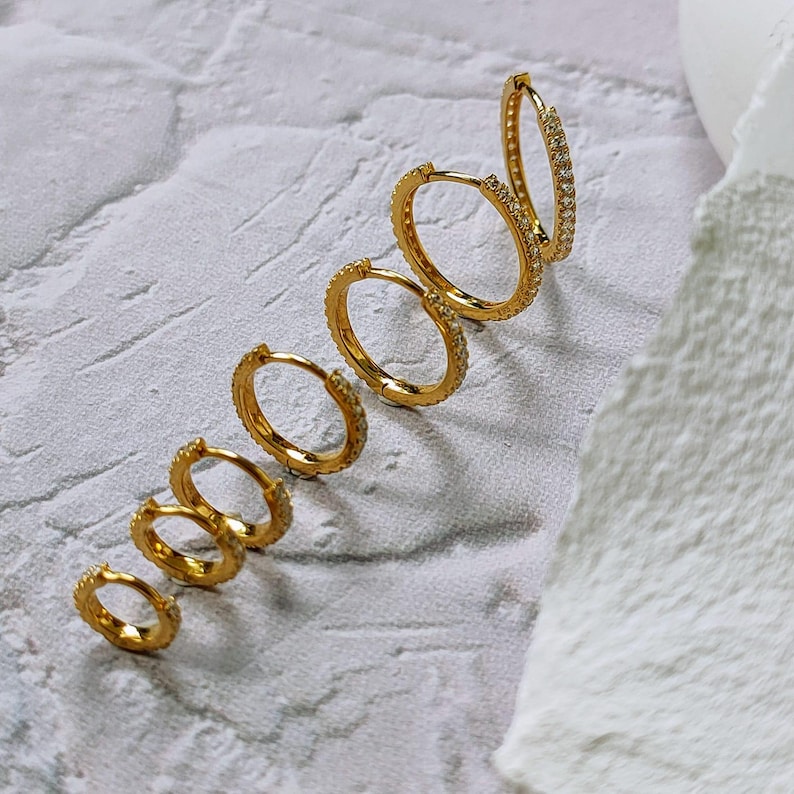 May include: A selection of gold-coloured hoop earrings in different sizes, each adorned with tiny, sparkling stones. The earrings are arranged diagonally on a textured, light-coloured surface, highlighting their design and detail.