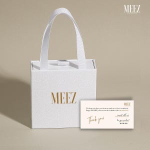 May include: A white gift bag with a handle, featuring the gold text "MEEZ". A small card with the text "Thank you!" and other text is placed in front of the bag.