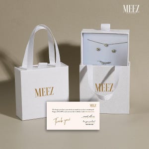 May include: White gift bags and a box with the gold text "MEEZ". The box is open, revealing a necklace, earrings, and a ring. A card with the text "Thank you!" is also present.