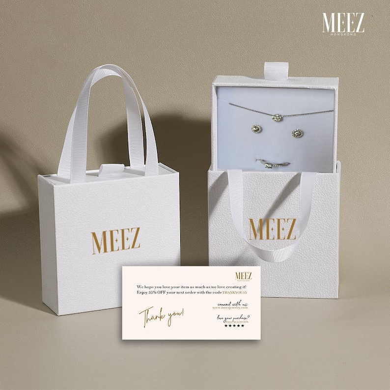May include: White gift bags and a box with the gold text "MEEZ". The box is open, revealing a necklace, earrings, and a ring. A card with the text "Thank you!" is also present.
