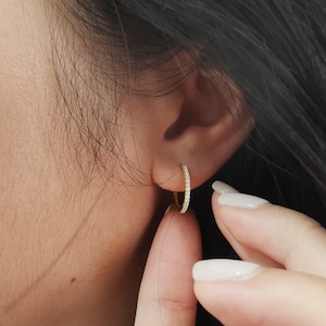 May include: A small, gold-colored hoop earring with a row of small, clear stones. The earring is being held by a person's finger. The earring is approximately 0.75 inches in diameter.