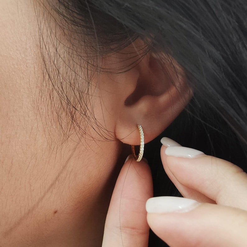 May include: A small, gold-colored hoop earring with a row of small, clear stones. The earring is being held by a person's finger. The earring is approximately 0.75 inches in diameter.