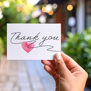 May include: A white card with the words "Thank you" in elegant black script, accompanied by a pink watercolor heart. The card is held against a blurred green background, suggesting an outdoor setting.
